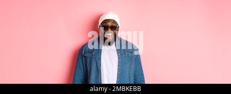Hipster afro-americano sorpreso e felice uomo in beanie e occhiali da sole, sorridendo e guardando la macchina fotografica, in piedi fiducioso su sfondo rosa Foto Stock