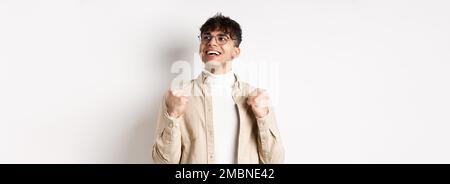 Il giovane fortunato dice sì con il viso sollevato, sorridendo e facendo pompa pugno, trionfando di successo, vincendo il premio, in piedi su sfondo bianco dopo Foto Stock