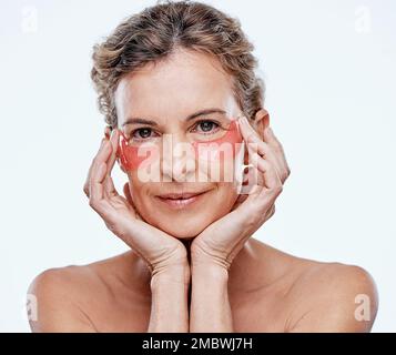 Provateli durante il vostro prossimo regime di auto-cura. una donna matura che indossa delle macchie di gel sotto gli occhi mentre si posa su uno sfondo bianco. Foto Stock