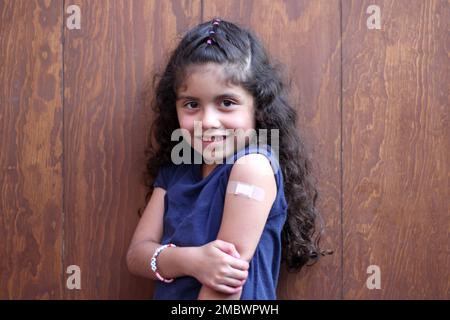 Piccola ragazza latina con capelli ricci felice mostra il suo braccio recentemente vaccinato contro Covid-19 nel nuovo normale per la pandemia di Coronavirus Foto Stock