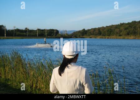 Un ingegnere ambientale che indossa un casco bianco utilizza un telefono cellulare per azionare un aeratore a turbina ad ossigeno in un laghetto. Concetto di acqua ed ecologia. Foto Stock