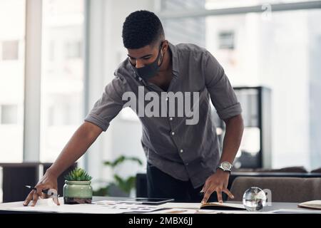 Riunendo tutte le sue bozze. un giovane designer che indossa una maschera facciale mentre passa attraverso la documentazione in un ufficio. Foto Stock
