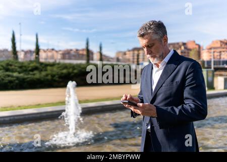 Serio uomo d'affari maturo in abbigliamento formale navigazione smartphone mentre in piedi sulla strada vicino fontana Foto Stock