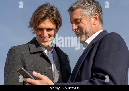 Uomini d'affari allegri in abiti formali sorridendo e navigando in smartphone mentre si levano in piedi insieme contro il cielo blu senza nuvole Foto Stock