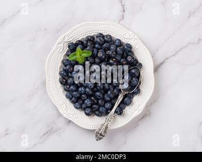 Mirtilli freschi e cucchiaio su un piatto bianco. Piatto pieno di frutti di bosco dolci maturi su fondo di marmo. Antiossidante naturale di mirtillo crudo. Vista dall'alto Foto Stock