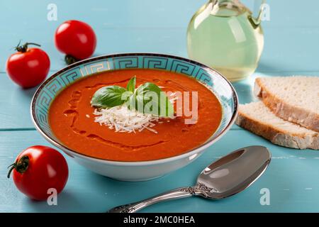 Ciotola per zuppa di pomodoro e cucchiaio su tavolo in legno turchese. Zuppa calda fatta in casa di pomodori arrosto con parmigiano e basilico verde. Vista frontale. Foto Stock