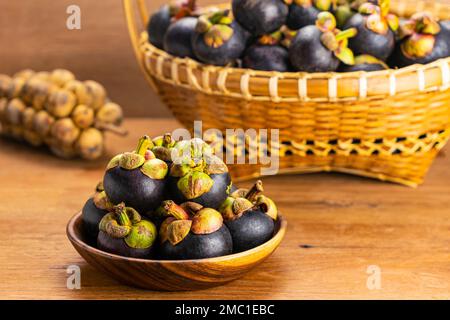 Mucchio di frutta fresca di mangosteo in piatto di legno e in cesto di bambù con mazzo di longkong su tavolo di legno Foto Stock