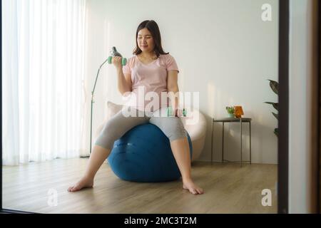 Bella donna asiatica incinta fare esercizio con manubri in soggiorno a casa, concetto sano Foto Stock