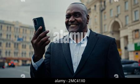 Sorridente afro-americano di mezza età etnica maschio uomo datore di lavoro imprenditore uomo d'affari in città strada all'aperto parlando telefono cellulare fotocamera parlare Foto Stock