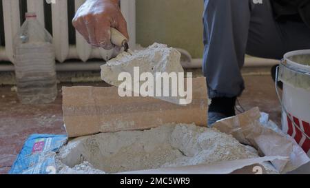 la mano del costruttore raccoglie lo stucco secco da una borsa di fabbrica e tiene una manciata di miscela sciolta su una cazzuola, preparando un mortaio per intonacare le pareti della stanza Foto Stock