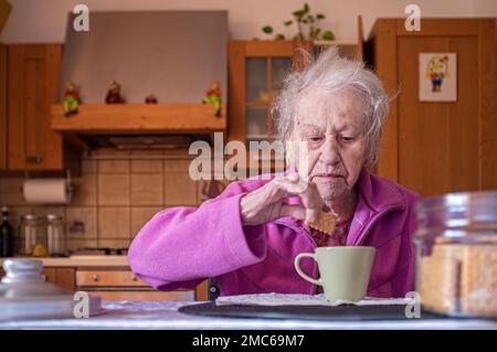 Donna molto anziana, sola nella sua cucina, immerge i biscotti in una tazza di caffè latte, durante la colazione Foto Stock