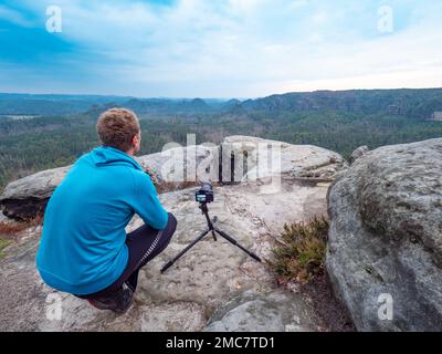 Zenzero uomo in felpa blu, si siede sui suoi talloni e aspetta che la fotocamera finisca l'esposizione della foto. Il fotografo lavora in natura. Foto Stock