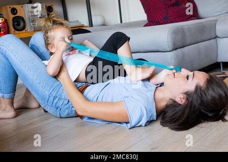 ragazza di 3 anni gioca seduta su sua madre, come se fosse una poltrona, con un nastro blu che tiene con i suoi piedi e le sue mani Foto Stock