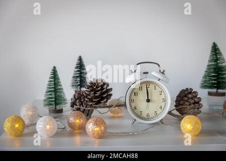 Una sveglia bianca, un puntatore a dodici, un piccolo albero di Natale verde artificiale e una ghirlanda su una cassettiera bianca. Spazio di copia. Biglietto di Natale. Foto Stock