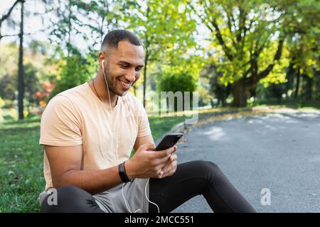 Uomo felice e soddisfatto dopo il jogging e la lezione di fitness seduti e utilizzando lo smartphone, uomo afroamericano in cuffie ascoltare podcast online, audiolibri e musica, digitando al telefono. Foto Stock