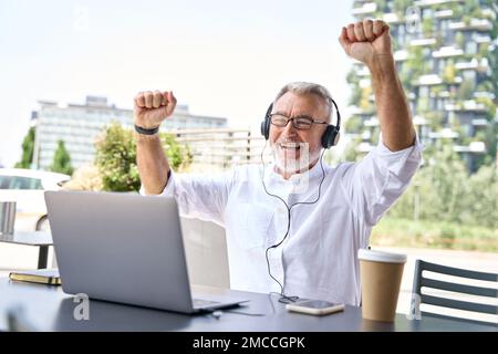 Un uomo d'affari anziano entusiasta che guarda al notebook che fa crescere i pugni vincendo online. Foto Stock