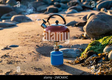 Stufa a gas turistica portatile con bollitore arancione sullo sfondo della natura sulla spiaggia. Campeggio cucina e tè. Foto Stock