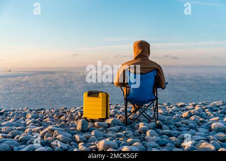 Uomo al barbuto in giacca marrone con valigia gialla che si rilassa da solo e si siede sulle sedie a sdraio sul mare all'alba. Concetto di stile di vita di viaggio Foto Stock