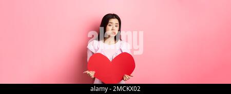 Concetto di San Valentino. Single teenage asian girl vuole innamorarsi, guardando triste e solitario alla macchina fotografica, sulking afflitto in festa degli amanti, tenendo grande Foto Stock