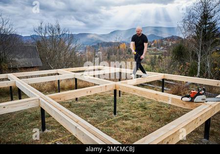 Uomo lavoratore costruire casa telaio in legno su fondazione palo in montagna. Falegname di misurazione tavole di legno. Concetto di Falegnameria. Foto Stock