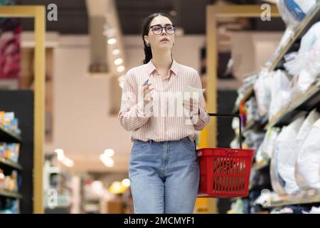 Giovane donna caucasica che indossa occhiali tiene il carrello rosso e camminare nel supermercato. Scaffali con cibo e navata in background. Concetto di shopping. Foto Stock