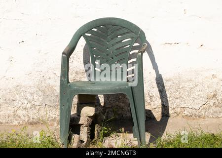 una vecchia sedia rotta di plastica si trova sullo sfondo di una parete bianca di una casa in cottage estivi al sole, una vecchia sedia è rotta Foto Stock