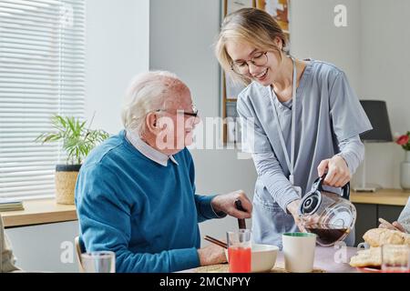 Giovane caregiver che versa il caffè dalla teiera per l'uomo anziano mentre fa colazione al tavolo Foto Stock