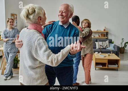 Felice coppia anziana ballare insieme in camera con altre persone e caregiver in background Foto Stock