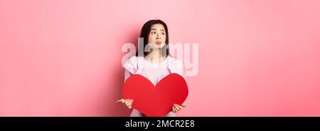 Concetto di San Valentino. Solitario adolescente asiatica ragazza sognando di amore, sentendosi triste e solitario in giornata degli amanti, guardando da parte con pietà, tenendo grande rosso Foto Stock