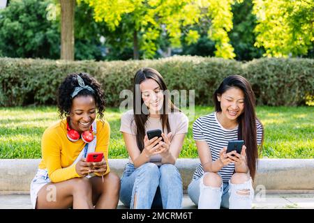 Adolescente donna amici che utilizzano il telefono cellulare insieme seduti all'aperto Foto Stock