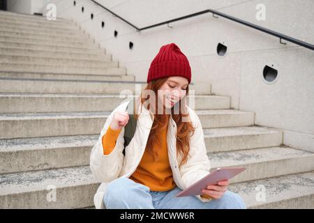 Giovane bella ragazza con lunghi capelli rossi, sorride, utilizza l'applicazione Internet, connettersi al wi-fi vicino al campus, siede sulle scale con tablet digitale Foto Stock