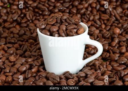 Una tazza bianca per espresso piena di chicchi di caffè interi si trova in un mucchio di chicchi di caffè tostati Foto Stock