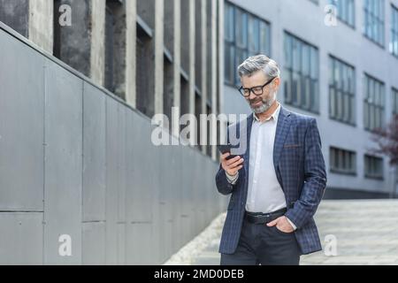 Boss dai capelli grigi di successo che cammina all'esterno dell'edificio dell'ufficio, uomo d'affari maturo che usa il telefono, sorridendo e leggendo le notizie che scrivono il messaggio, uomo dai capelli grigi in vestito di affari e negli occhiali. Foto Stock