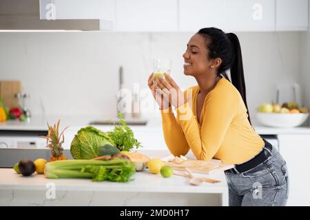 Sorridente giovane afro-americana bere un bicchiere di frullato al tavolo con verdure biologiche, gustare la dieta Foto Stock