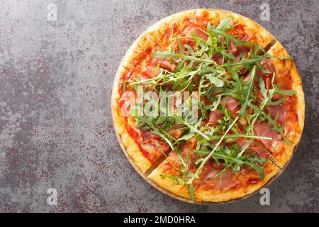 Pizza calda con prosciutto e rucola in primo piano su un asse di legno sul tavolo. Vista orizzontale dall'alto Foto Stock