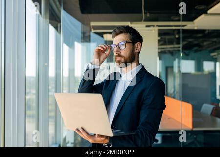 Serio uomo d'affari di mezza età negli occhiali che tengono il laptop e che guarda da parte, in piedi vicino alla finestra negli interni dell'ufficio Foto Stock