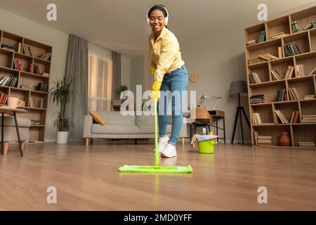 Giovane donna nera lavando pavimento in soggiorno utilizzando mop e ascoltando musica in cuffie, godendo di pulizia routine Foto Stock