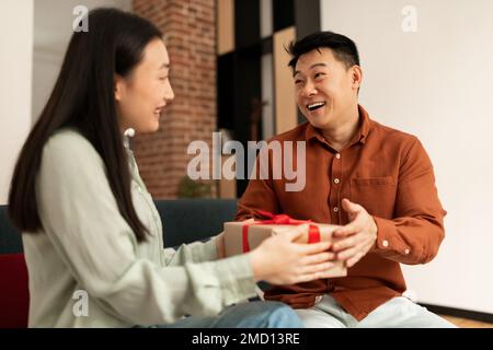 Amante giovane moglie asiatica che dà regalo al suo marito maturo felice, seduto sul divano a casa Foto Stock
