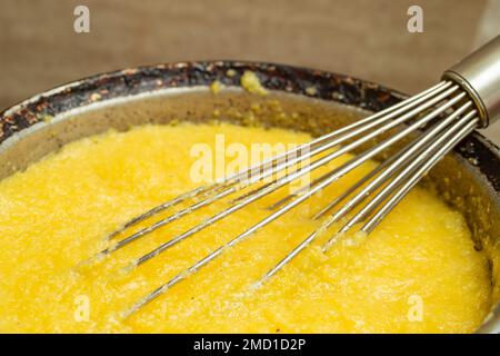 Far bollire la farina di mais in una pentola di ghisa, preparare mamaliga, cibo tradizionale rumeno, e mescolare con una frusta in acciaio inossidabile, fuoco morbido primo piano Foto Stock