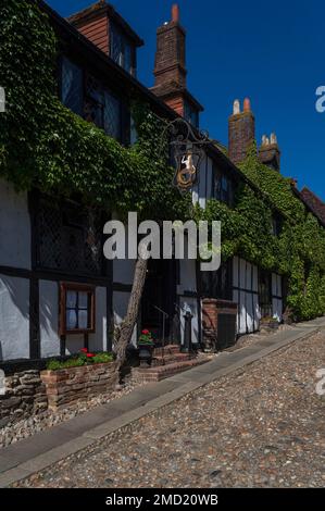 La glicine fiorisce attraverso la parte anteriore incorniciata in legno dell'antica Mermaid Inn in strada acciottolata Mermaid Street a Rye in East Sussex, Inghilterra, Regno Unito. L'insegna della locanda, che oscilla da elaborati lavori in ferro battuto, presenta una sirena dai capelli dorati con una coda dorata. Foto Stock