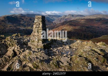 Trig Point a Loughrigg cadde sostenuto da Great Langdale & The Langdale Pikes in autunno, Lake District National Park, Cumbria, Inghilterra, Regno Unito Foto Stock