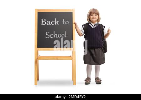 Allievo femminile in una divisa scolastica che tiene un gesso e che si trova accanto a una lavagna con testo di ritorno a scuola isolato su sfondo bianco Foto Stock