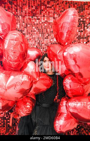 giovane donna in stativo nero con palloncini a forma di cuore su sfondo rosso Foto Stock