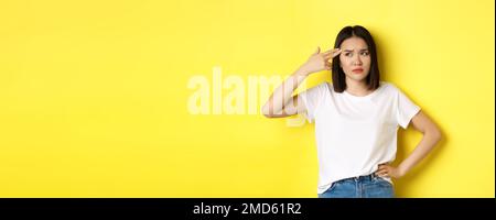 Donna che mi mostra uccidere prego gesto, sparando in testa con la pistola dito dalla noia, in piedi sconvolto su sfondo giallo Foto Stock