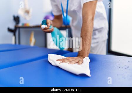 Giovane uomo ispanico che indossa fisioterapista uniforme disinfezione tavolo da massaggio alla clinica di riabilitazione Foto Stock