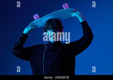 Adolescente maschio con cuffie ascoltare musica e ballare e cantare con skateboard in mano sulla sua testa, stile di vita hipster, sfondo blu Foto Stock