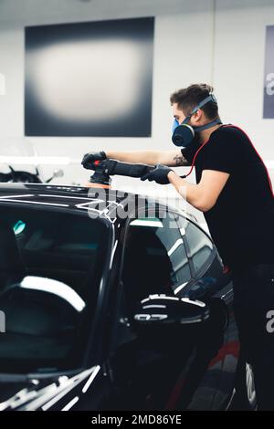 Uomo lucidando la vernice dell'automobile. Scatto verticale. Persona professionista che si dettaglio auto in maschera di protezione del viso utilizzando la lucidatrice orbitale. Foto di alta qualità Foto Stock