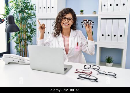 Giovane donna bruna che tiene occhiali da optometria sorridente felice di indicare mano e dito di lato Foto Stock