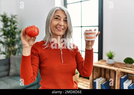 Donna di mezza età dai capelli grigi sorridente sicura che tiene la mela rossa e la dentiera a casa Foto Stock