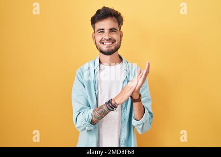 Giovane uomo ispanico con tatuaggi in piedi su sfondo giallo che batte e applaude felice e gioioso, sorridendo mani orgogliose insieme Foto Stock